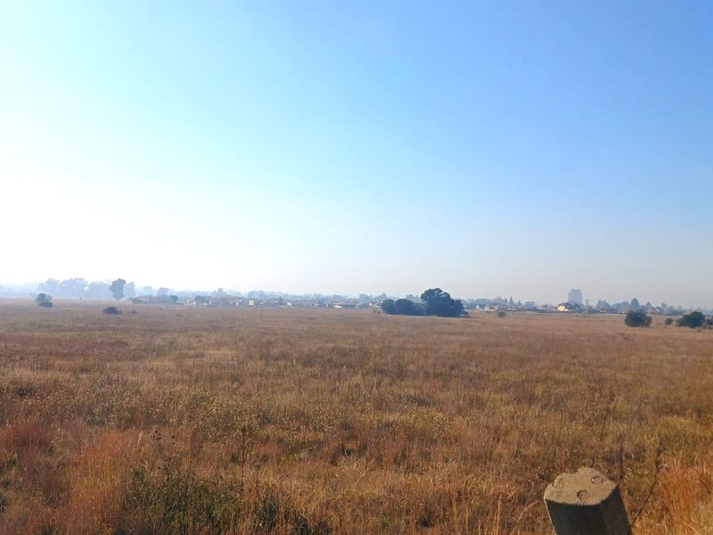 A wide-angle view of a vacant residential land plot featuring dry grass and distant neighborhood scenery.