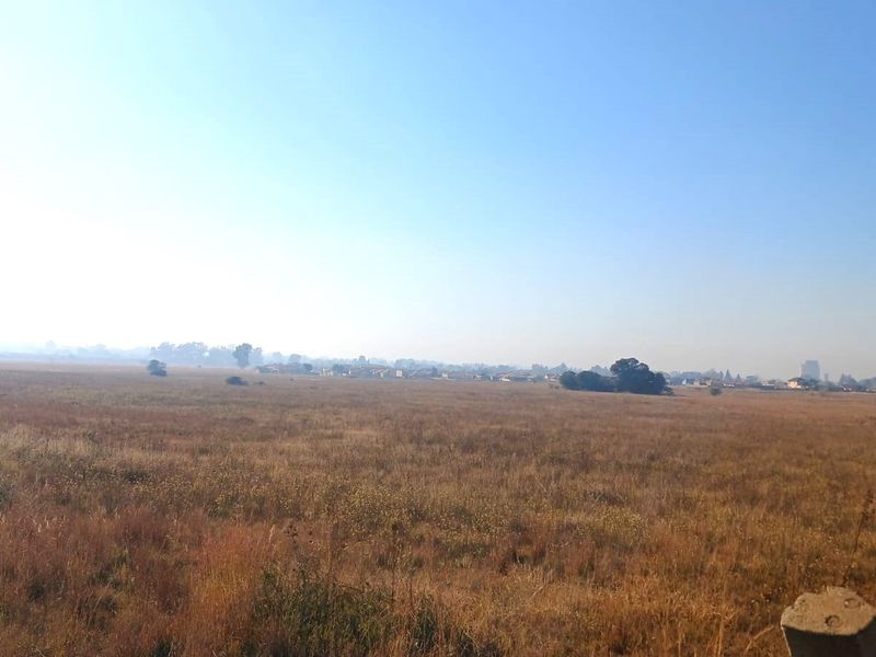 A wide-angle view of an expansive, undeveloped residential land plot featuring natural grassland with distant suburban structures visible on the horizon.