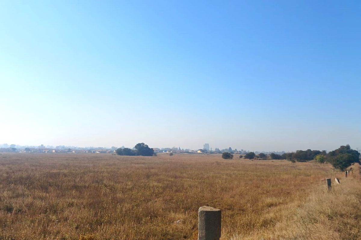 A wide-angle view of a vacant residential plot featuring dry grass, a dirt access road, and distant neighborhood scenery.