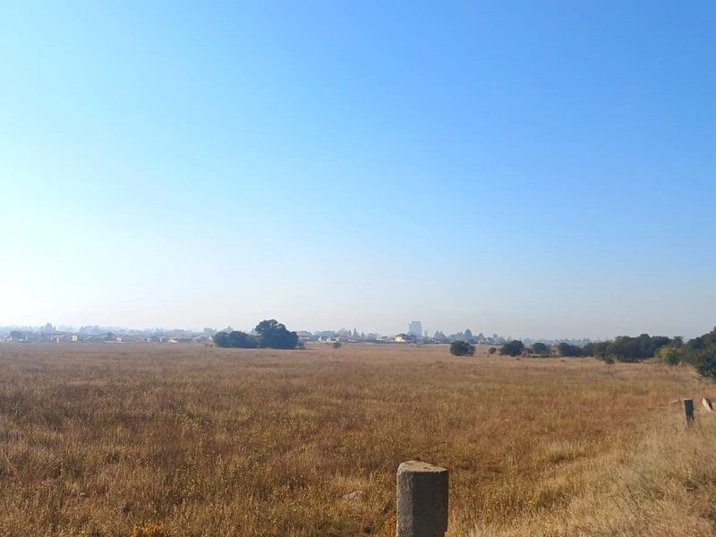 A wide-angle view of a vacant residential plot featuring dry grass, a dirt access road, and distant neighborhood scenery.