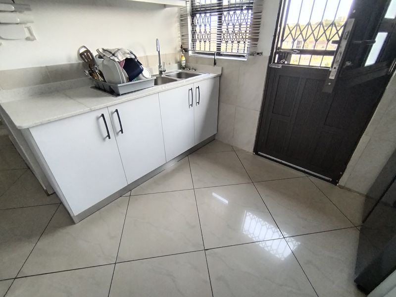 A functional kitchen area featuring white cabinetry, a stainless steel double sink, tiled flooring, and a security gate leading to the exterior.