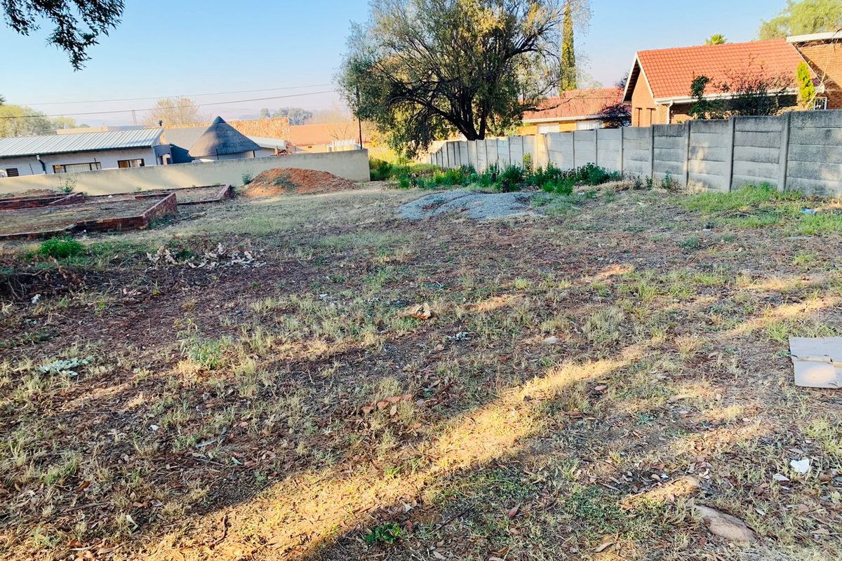 A vacant residential plot featuring a precast concrete boundary wall, uneven terrain, and remnants of brick foundations, offering potential for development.