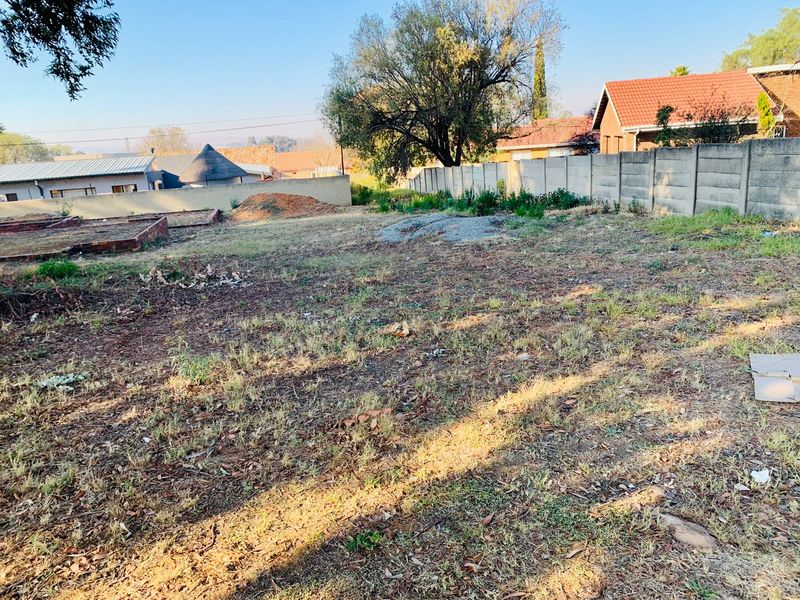 A vacant residential plot featuring a precast concrete boundary wall, uneven terrain, and remnants of brick foundations, offering potential for development.