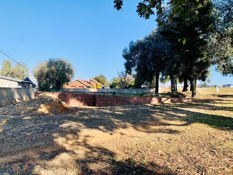 A vacant residential plot featuring a brick retaining wall, mature trees, and established boundary fencing, offering a clear space for future development.