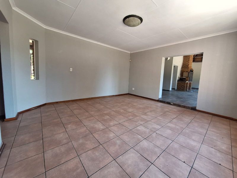 An empty, neutrally painted living room featuring tiled flooring and a coved ceiling, with a view through to an adjacent room with a brick fireplace.