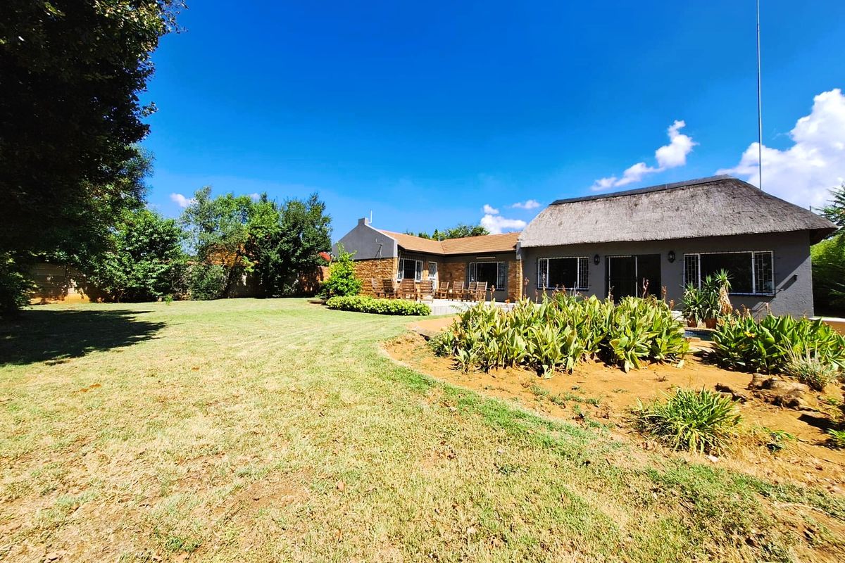 Spacious rear garden featuring a well-maintained lawn, landscaped garden beds, and a paved patio area adjacent to the house with a traditional thatched roof.