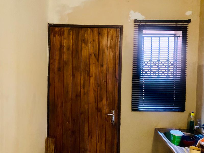 A small kitchen area featuring a stainless steel sink, wooden door, and window with venetian blinds, currently requiring wall finishing and painting.