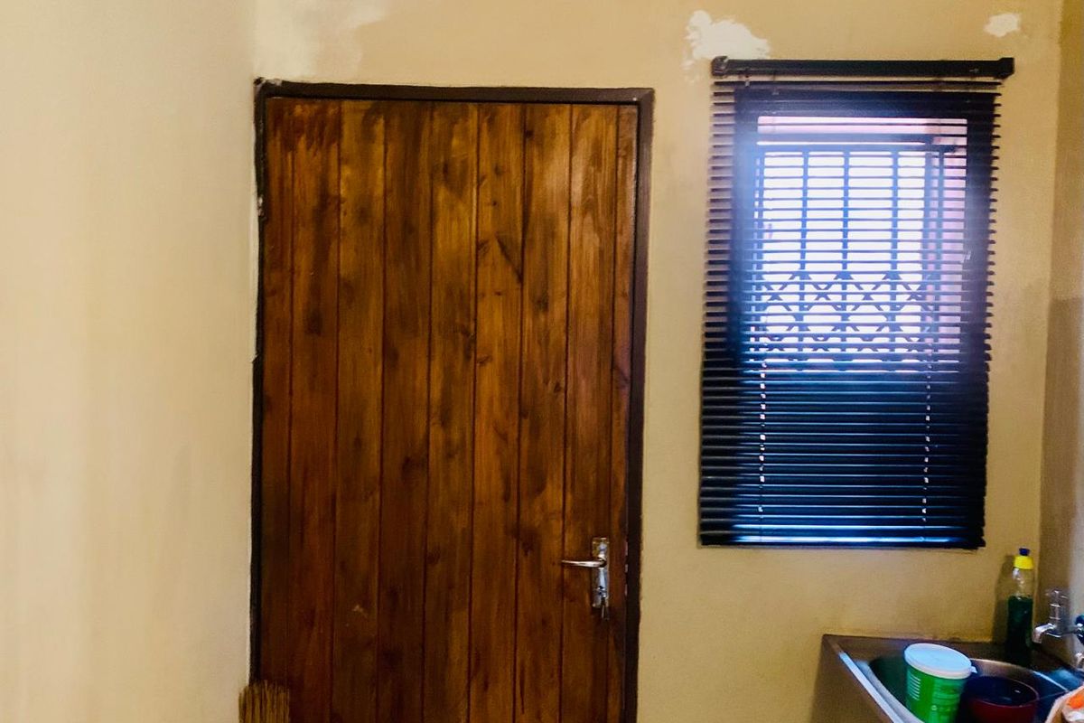 A small kitchen area featuring a stainless steel sink, wooden door, and window with venetian blinds, currently requiring wall finishing and painting.