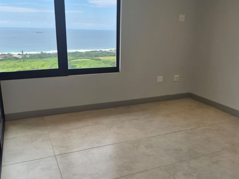 A bright, empty bedroom featuring large format floor tiles, modern black aluminium window frames, and a scenic coastal view.