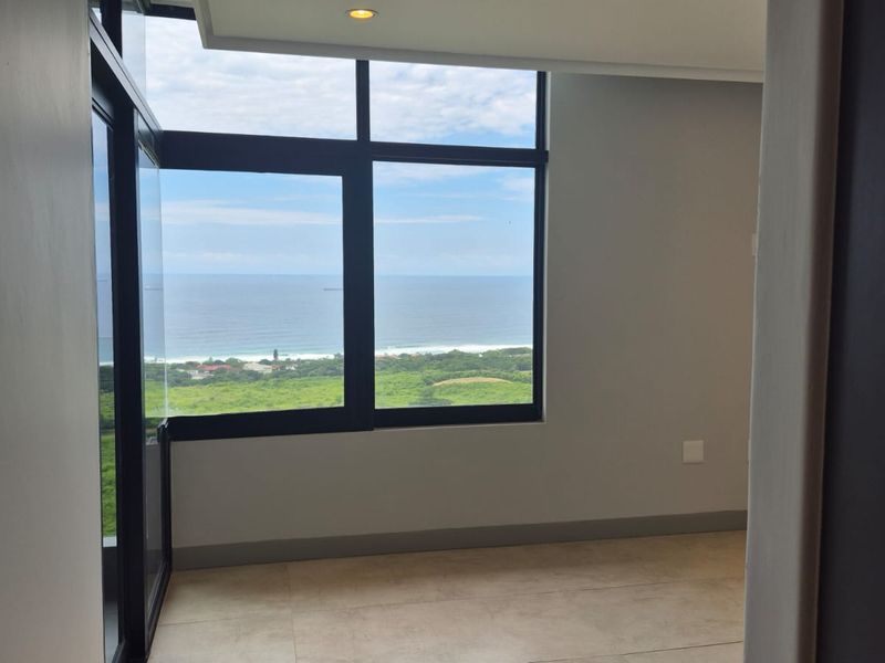 A bright, empty bedroom featuring large windows with expansive ocean and coastal views, neutral tiled flooring, and modern recessed lighting.