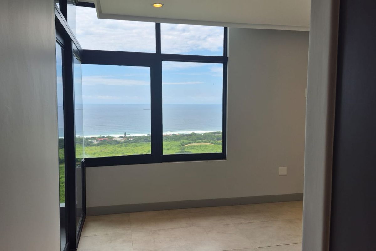 A bright, empty bedroom featuring large windows with expansive ocean and coastal views, neutral tiled flooring, and modern recessed lighting.