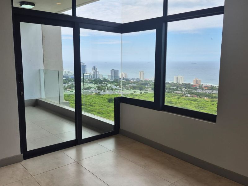 Bright, empty living area featuring floor-to-ceiling windows, tiled flooring, and direct access to a balcony with expansive coastal and city views.