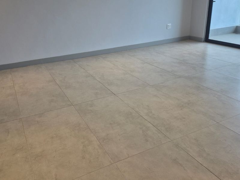 A bright, empty living area featuring modern large-format floor tiles, neutral walls, and a sliding glass door leading to an outdoor balcony.