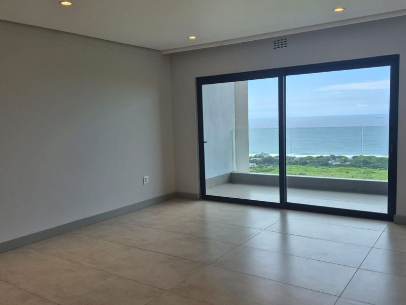 A bright, empty living area featuring large sliding glass doors that open onto a balcony with expansive ocean views.