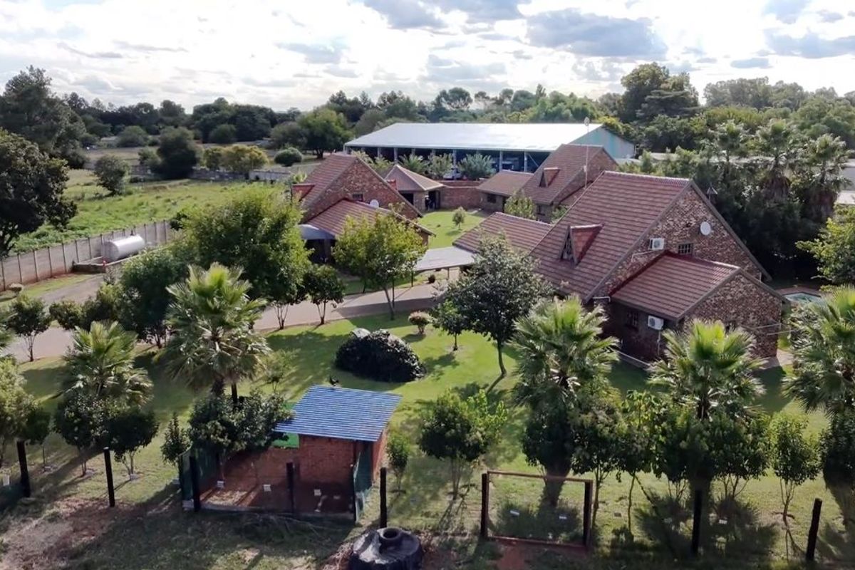 Aerial view of a farm property featuring multiple brick buildings with tiled roofs, surrounded by established gardens and mature trees.