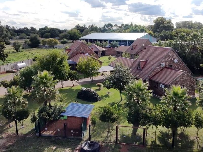 Aerial view of a farm property featuring multiple brick buildings with tiled roofs, surrounded by established gardens and mature trees.