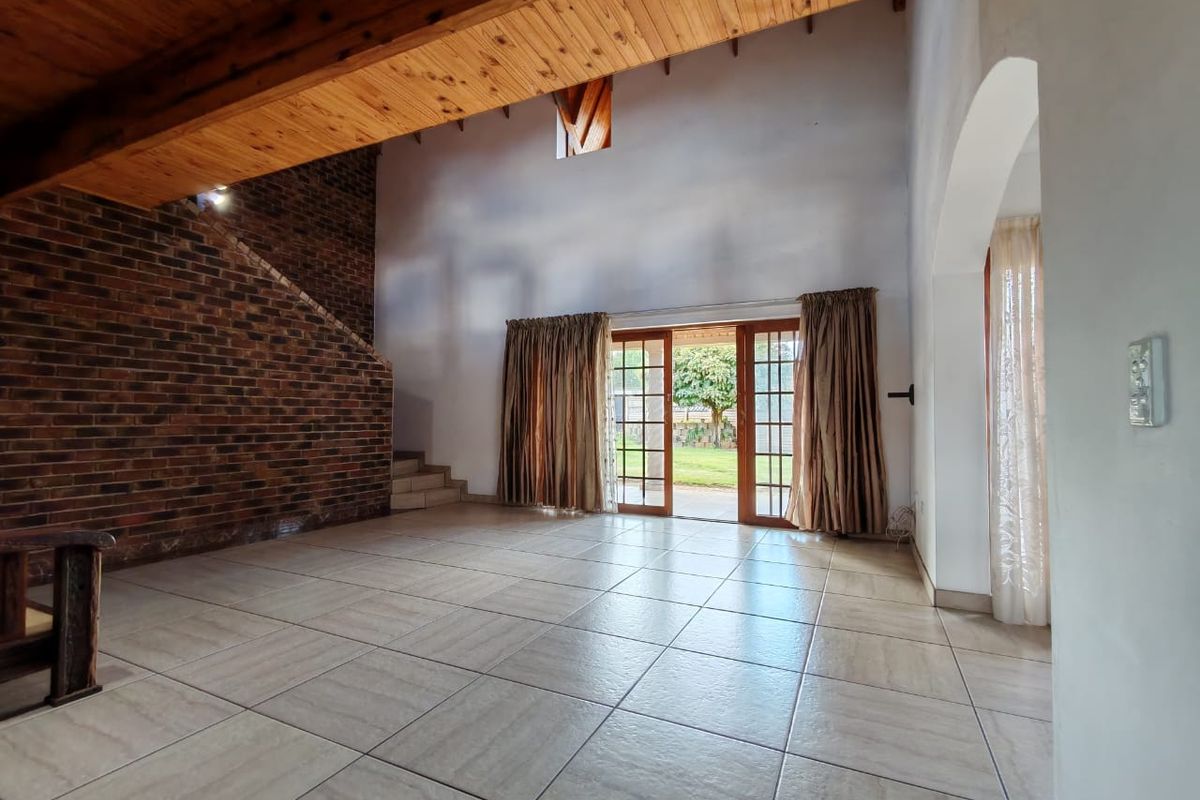 Spacious living area featuring high ceilings with exposed timber beams, an exposed brick wall, tiled flooring, and direct access to the garden through glass doors.