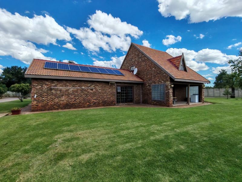 Rear view of a facebrick farm-style residence featuring a terracotta tiled roof, integrated solar panels, and a covered patio area set within a lawn.