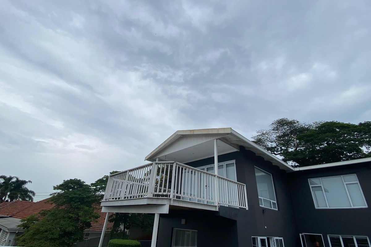 A rear view of the house featuring a raised balcony with white balustrades, large windows, and dark exterior paint under a cloudy sky.