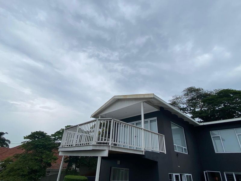 A rear view of the house featuring a raised balcony with white balustrades, large windows, and dark exterior paint under a cloudy sky.
