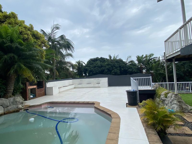 Outdoor pool area featuring a swimming pool, built-in brick braai, concrete seating, and raised deck with railing, set within a landscaped garden.
