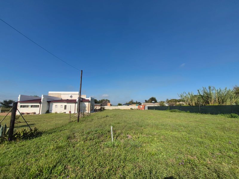 A wide-angle view of a vacant residential plot featuring a grass lawn, perimeter fencing, and an adjacent building structure.