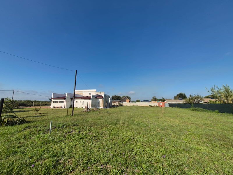 A spacious residential plot featuring a large grass lawn, perimeter fencing, and a concrete boundary wall, with an existing structure visible in the background.