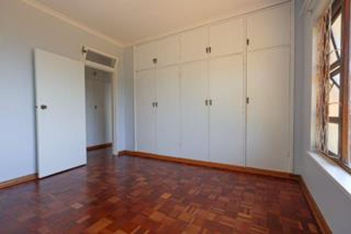 A bright bedroom featuring classic parquet flooring, extensive built-in floor-to-ceiling cabinetry, and a large window with security bars.
