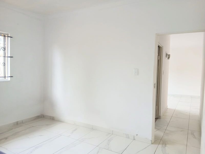 A bright, empty bedroom featuring white walls, marble-look tiled flooring, and a window with security bars, leading into an adjacent hallway.