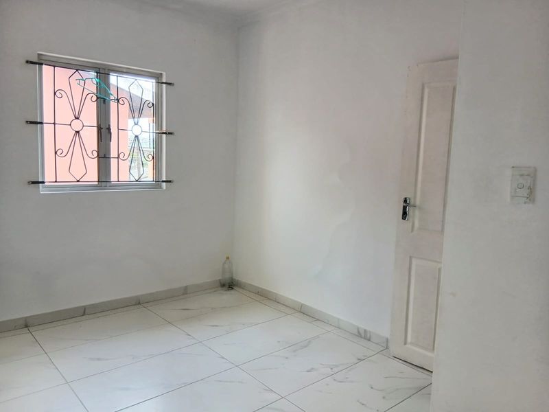 A vacant bedroom featuring white walls, tiled flooring, and a window with security bars. Visible water staining on the wall requires attention.