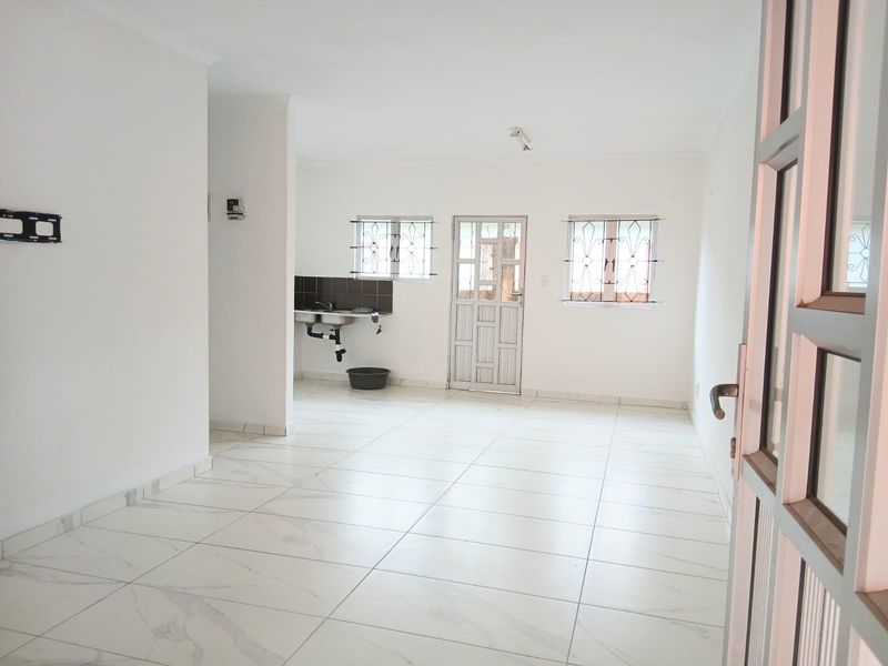 An open-plan living and kitchen area featuring white tiled flooring, security bars on windows, and a basic kitchen sink area.
