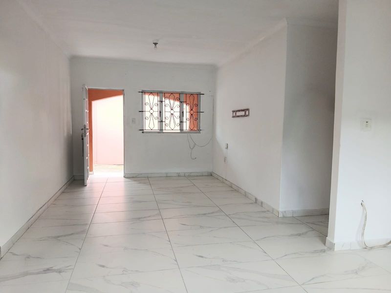 An empty, open-plan living area featuring white tiled flooring, neutral walls, and a window fitted with security bars.