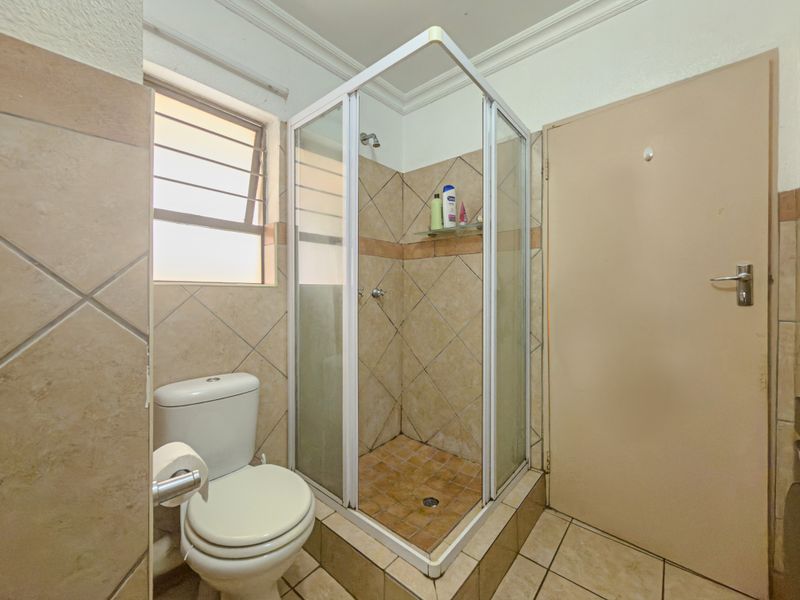 A compact bathroom featuring a corner shower enclosure, toilet, and neutral wall and floor tiling with a louver window for ventilation.
