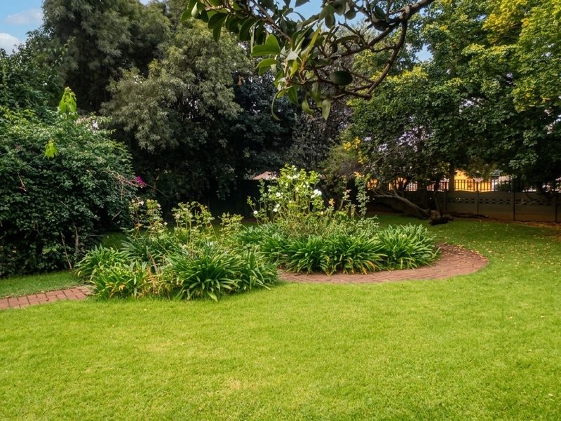 A well-maintained garden featuring a brick paved pathway, mature trees, and established garden beds within a private boundary wall setting.