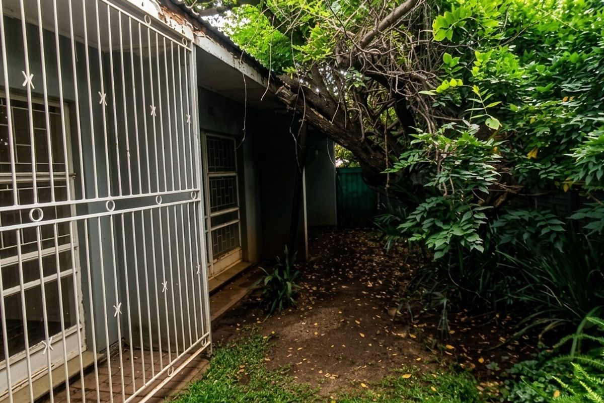 A view of the rear exterior of the house featuring security bars on windows and doors, with overgrown garden foliage adjacent to the building structure.