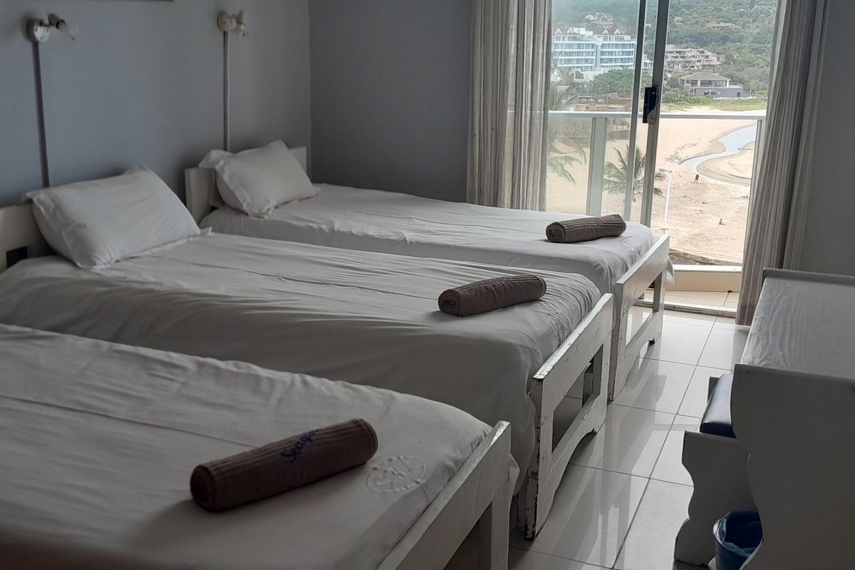 A bedroom featuring three single beds, tiled flooring, and a sliding glass door leading to a balcony with coastal views.