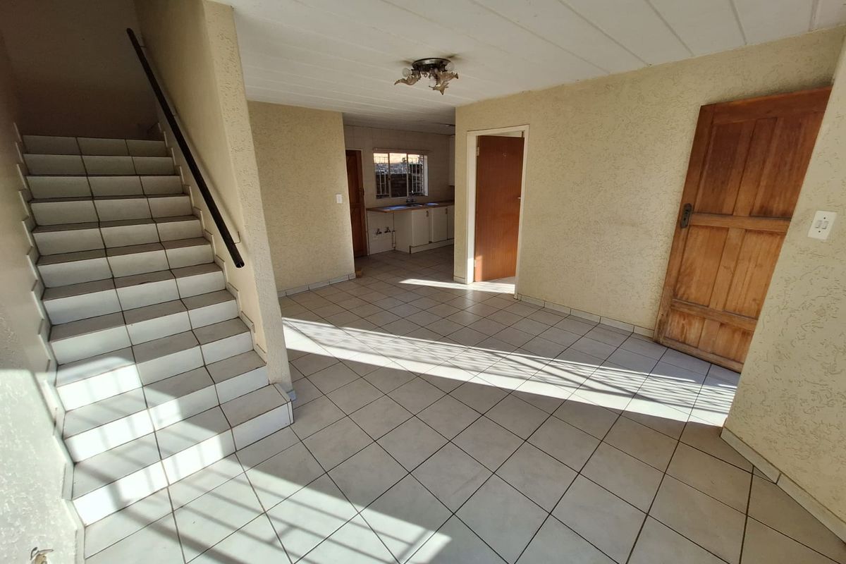 An empty, open-plan living area featuring tiled flooring, a staircase with a handrail, and direct access to an adjacent kitchen space.