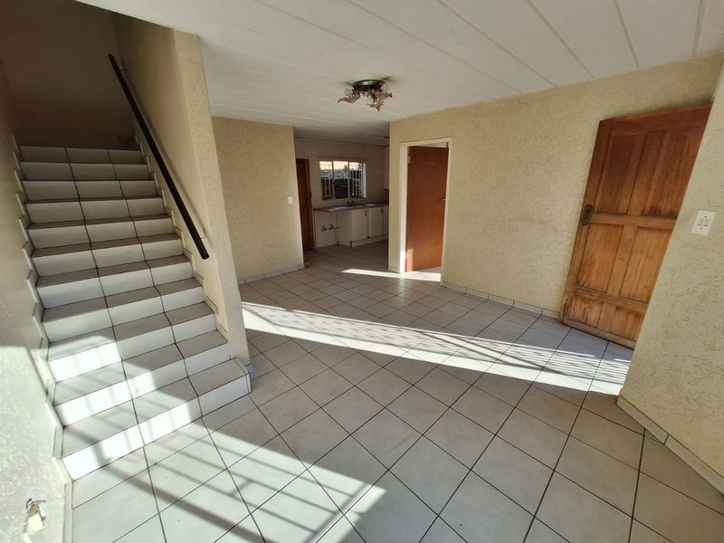An empty, open-plan living area featuring tiled flooring, a staircase with a handrail, and direct access to an adjacent kitchen space.