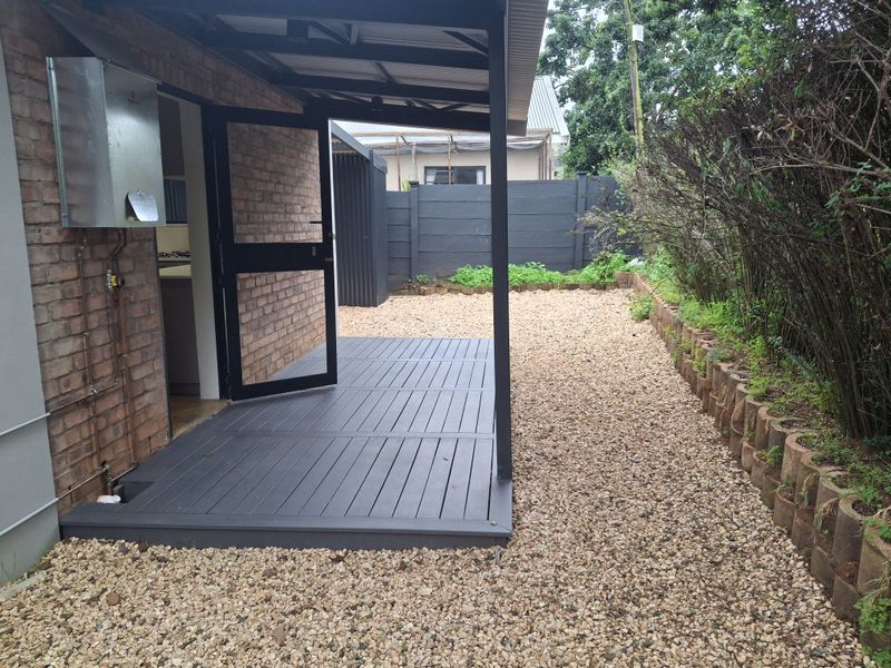 A neat covered patio area featuring composite decking, exposed brick walls, and a gas geyser, leading out to a low-maintenance gravel garden.