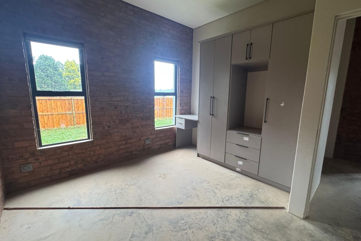 A spacious bedroom featuring an exposed brick wall, built-in cupboards with an integrated desk, and large windows overlooking the garden.