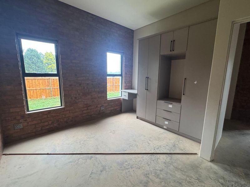 A spacious bedroom featuring an exposed brick wall, built-in cupboards with an integrated desk, and large windows overlooking the garden.
