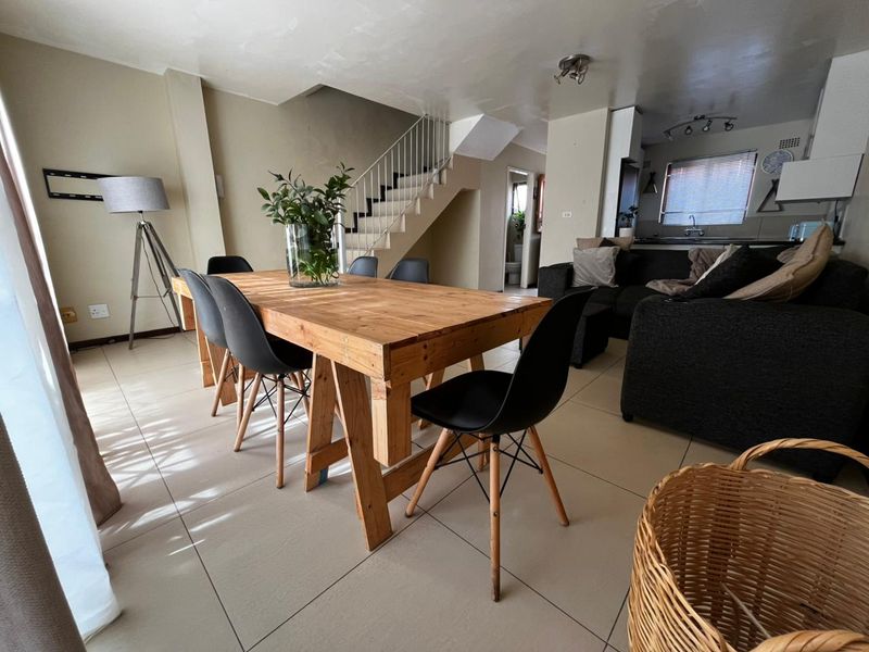 Open-plan living and dining area featuring tiled floors, a staircase with metal railing, and a functional kitchen space in the background.