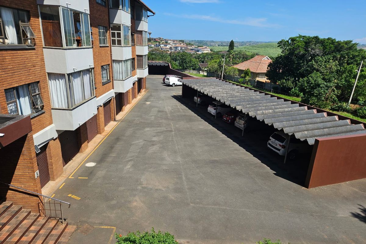 An elevated view of the apartment complex exterior, featuring a paved parking area with covered carports and brick building facades.