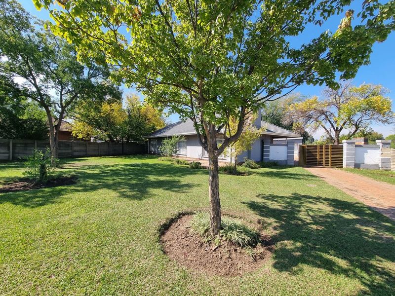 A spacious front garden featuring a well-maintained lawn, mature trees, a concrete driveway, and secure perimeter walling with a metal gate.