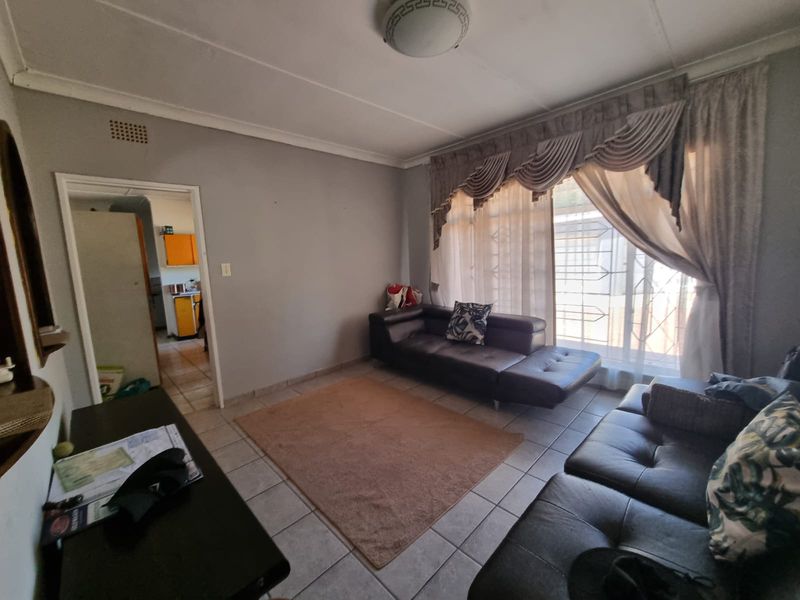 A living room featuring tiled flooring, a security gate on the window, and access to an adjacent kitchen area.