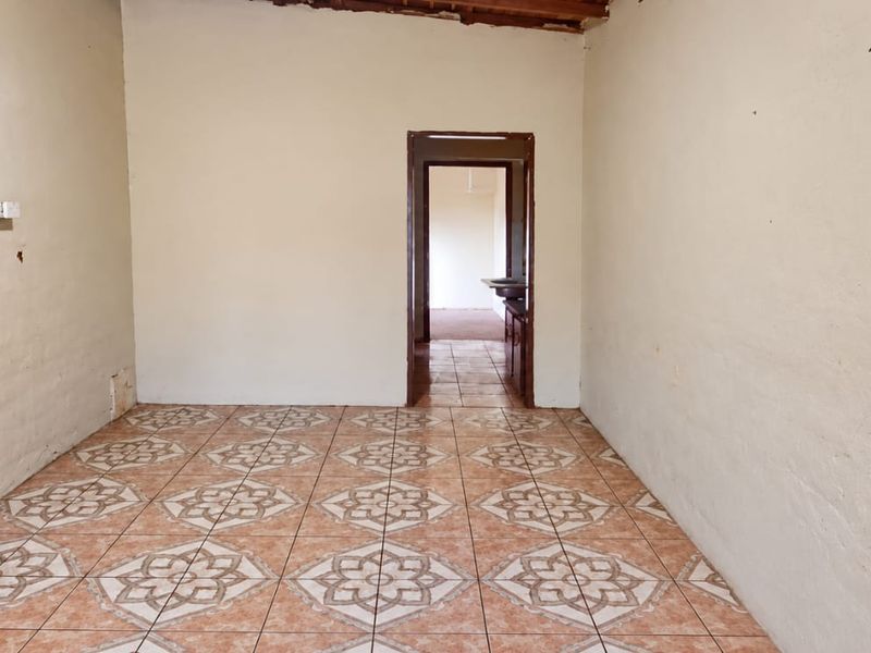 An empty living room featuring patterned ceramic floor tiles and exposed timber ceiling beams, requiring cosmetic updates and wall repairs.
