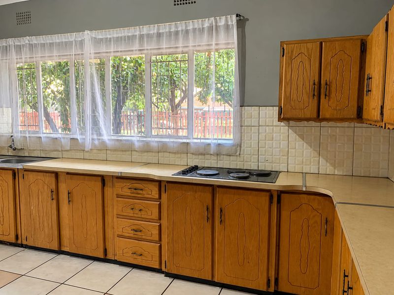 A functional kitchen featuring traditional wooden cabinetry, laminate countertops, a built-in electric hob, and large windows with security bars.