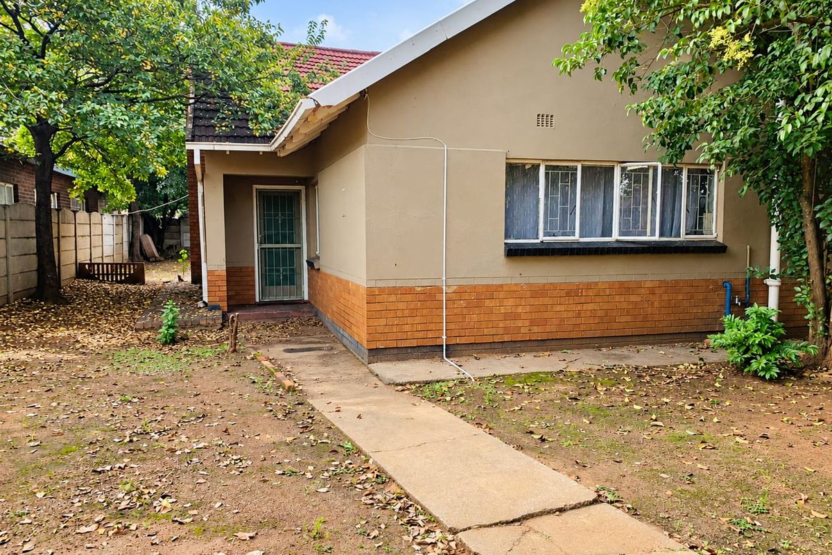 Rear view of a single-story brick house featuring a concrete walkway, security gate, and tiled roof, set within a garden area.