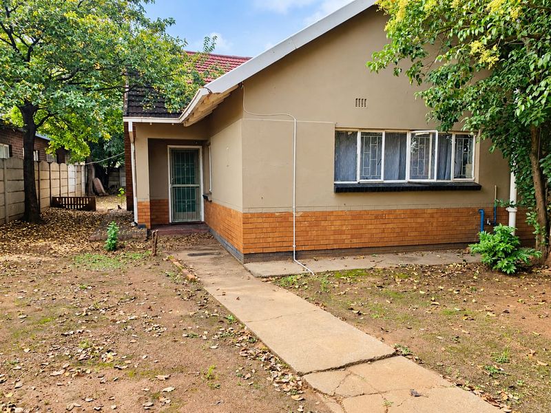Rear view of a single-story brick house featuring a concrete walkway, security gate, and tiled roof, set within a garden area.