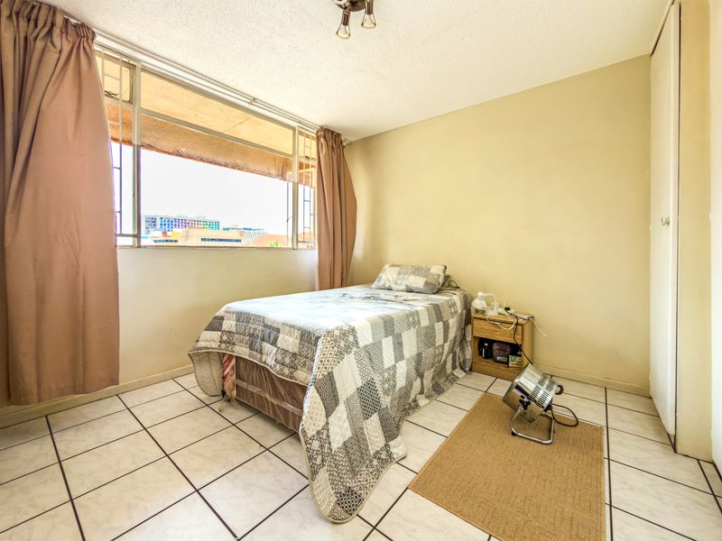 A bedroom featuring tiled flooring, a built-in cupboard, and a large window with security bars, offering natural light and city views.